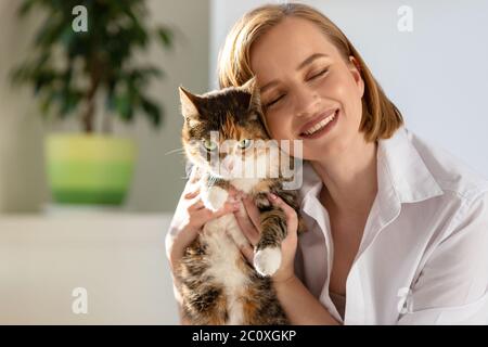 Nahaufnahme der lächelnden Frau in weißem Hemd umarmt und umarmt mit Zärtlichkeit und Liebe Hauskatze in zu Hause. Liebe zu den Tieren, Haustiere Konzept Stockfoto
