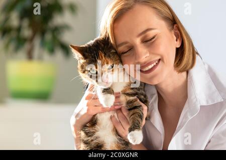 Nahaufnahme der lächelnden Frau in weißem Hemd umarmt und umarmt mit Zärtlichkeit und Liebe Hauskatze in zu Hause. Liebe zu den Tieren, Haustiere Konzept Stockfoto