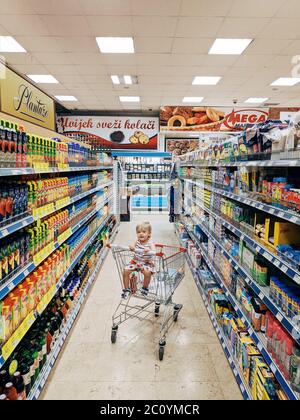 Budva, Montenegro - 10. juni 2020: Ein kleines Kind sitzt in einem Trolley vom Supermarkt. In der Abteilung mit Säften und Haushaltswaren. Stockfoto