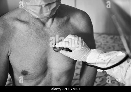 Ausschnitt aus der Hand des Arztes in einem Schutzanzug, der ältere männliche Patienten zu Hause mit Phonendoskop untersucht, Brustkorb überprüft, Schwarz-Weiß-Bild. COVID-19 Konzept Stockfoto