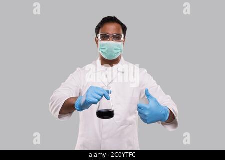 Männlich Arzt Holding Flasche mit Colorfull Flüssigkeit zeigt Daumen nach oben Tragen von medizinischen Maske, Handschuhe und Brille isoliert. Wissenschaft, Medizin, Virenkonzept Stockfoto