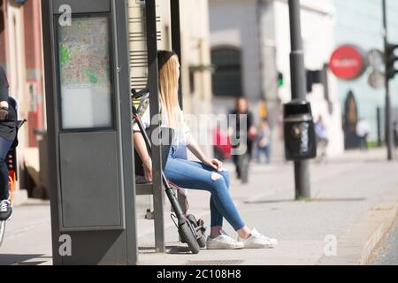 Casual kaukasischen Teenager Pendler mit modernen faltbaren städtischen Elektroroller sitzt auf einer Bushaltestelle Bank warten auf Metro City Bus. Mobilität in der Stadt Stockfoto