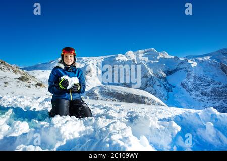 Lächelndes Teenager-Mädchen in blauem Outfit Maske und Helm zeigen Liebe Konzept hält Schnee Form im Herzen auf Knien mit Berggipfel auf dem Hintergrund stehen Stockfoto