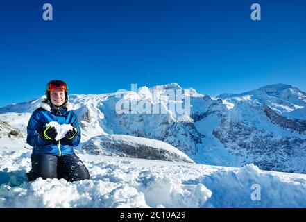 Happy Teenager-Mädchen halten Herz geformten Schnee zeigt Liebe Konzept auf ihren Knien mit Bergspitzen auf dem Hintergrund stehen Stockfoto