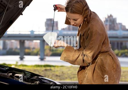 Selbstbewusste kaukasische Frau Fahrer Überprüfung Ölstand im Auto Motor Messstab, Stadtbild auf Hintergrund Stockfoto