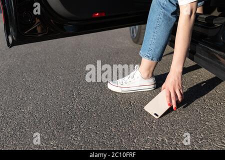 Frau stieg in ein Auto und ließ Smartphone auf Asphalt fallen, brach das Glas auf der Rückseite und nahm ein kaputtes Telefon vom Boden. Stockfoto