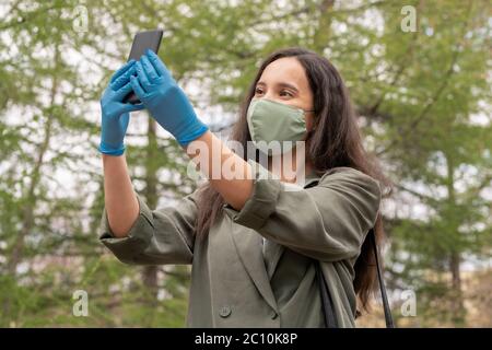 Schöne junge Frau in Stoffmaske und Latexhandschuhe mit Smartphone während der Aufnahme Selfie in Maske im Freien Stockfoto
