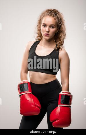 Blonde fit Mädchen in schwarzem Trainingsanzug und roten Boxhandschuhe stehen vor der Kamera isoliert bereit für das Training im Sportverein oder Fitnessstudio Stockfoto