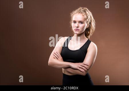 Serious Young fit weibliche Athletin mit langen blonden lockigen Haar Crossing ihre Arme auf der Brust, während sie vor braunem Hintergrund Stockfoto