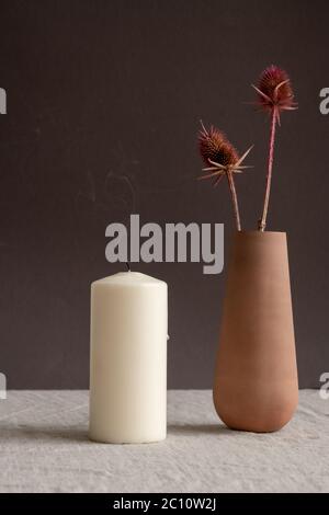Ausgeblasenes aromatisches Kerzenlicht und handgemachte braune Tonvase oder Krug mit zwei trockenen Wildblumen, die auf dem Tisch gegen die schwarze Wand stehen Stockfoto