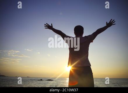 Silhouette eines Mannes, der seine Hände hebt oder die Arme am Strand bei Sonnenuntergang öffnet Stockfoto