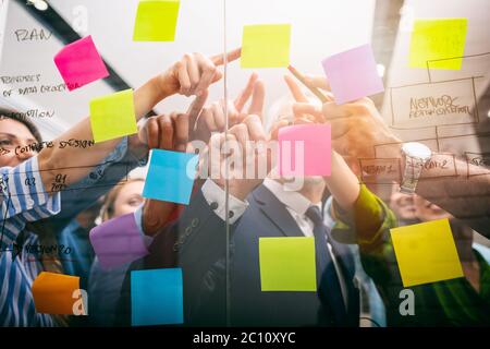 Das Team von Leuten bei der Arbeit im Büro zeigt mit der Hand einen Post-it auf dem Glas Stockfoto