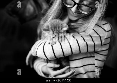 Ein junges Mädchen mit Brille hält zu Hause ein kleines, zwei Monate altes schottisches, gerades Kätzchen in den Armen. Schwarzweiß Foto mittlerer Grundriss Stockfoto