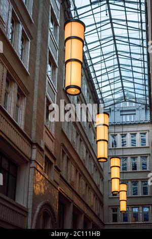Blick auf glühende mit gelbem Licht große, zylindrische, Pendelleuchten in einem Durchgang mit einem Glasdach. Stimmungsvolle Beleuchtung mit warmweißem Licht. Stockfoto