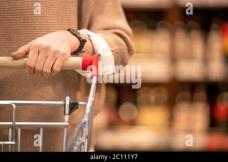 Hand der reifen weiblichen Kunden der zeitgenössischen Supermarkt schieben Warenkorb mit Waren beim Besuch einer der Abteilungen Stockfoto