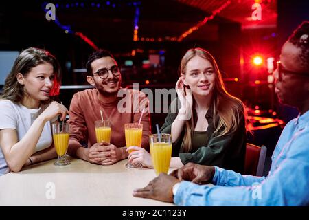 Zwei Mädchen aus kaukasischer Ethnie und gemischter Rasse, die ihrem afrikanischen Freund zuhören, der ihnen von sich selbst erzählt, indem sie in der Bar ein Glas Saft trinken Stockfoto