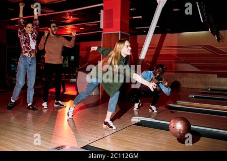 African guy Foto von jungen Frau werfen Ball auf Bowling-Bahn, während ihre begeisterten Freunde in der Nähe von durch Anheben der Hände durng Spiel Stockfoto