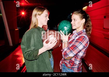 Zwei glückliche Mädchen in Casualwear halten Bälle während etwas auf dem Hintergrund der Bowling-Bahnen im dunklen Entertainment-Club diskutieren Stockfoto