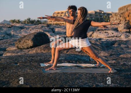 Paar, die Yoga bei Sonnenaufgang am Strand mit Blick auf das Meer praktizieren Stockfoto