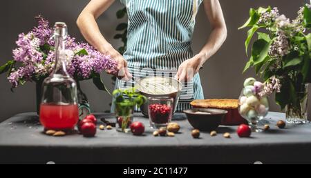 Kochen hausgemachte Cranberry Pie. Eine weibliche Konditorin peitscht einen Schneebesen mit saurer Sahne. Tisch mit hellen Zutaten im Frühlingsstil mit Blumen Stockfoto