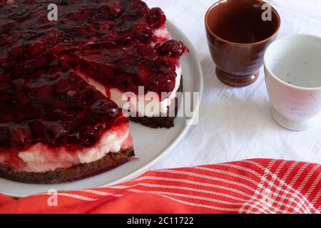 Hausgemachte Torte in Scheiben geschnitten, mit Mascarpone-Käsecreme, Gelee und frischen Kirschen auf einer weißen Keramikplatte auf einem weißen und roten Backgroun Stockfoto
