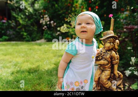 Lächelndes kleines Mädchen in einem türkisfarbenen Tuch, das in der Nähe einer kleinen Statue von Jungen und Mädchen steht. Kleines Mädchen Porträt Stockfoto