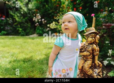 Lächelndes kleines Mädchen in einem türkisfarbenen Tuch, das in der Nähe einer kleinen Statue steht. Kleines Mädchen Porträt Stockfoto