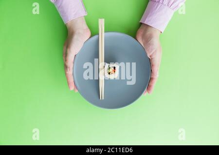 Flache Lay von weiblichen Händen halten Platte mit Essstäbchen und eine Sushi-Rolle isoliert auf grünem Hintergrund. Stockfoto