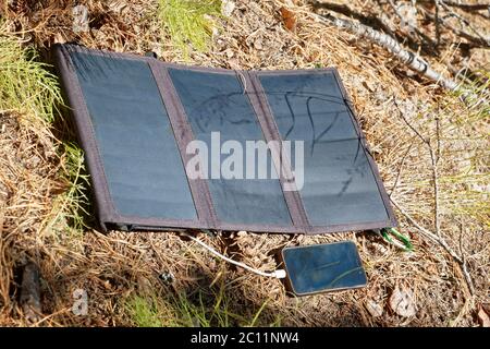 Das faltbare Solarmodul liegt auf der Oberfläche des Waldbodens und Gebühren das Smartphone. Komfort für Reisen und lange Spaziergänge vom traditio Stockfoto