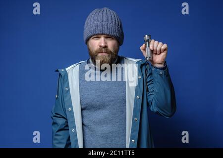 Serious bärtigen Mann in einem Strickmütze und einer wasserdichten Jacke hält eine Taschenlampe in den Händen, auf einem blauen Hintergrund. Stockfoto