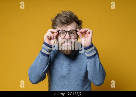 Der zottelige, bärtige Mann in einem alten Pullover mit einem seltsamen Gesichtsausdruck hält eine Brille in den Händen und versucht, etwas zu sehen. Stockfoto