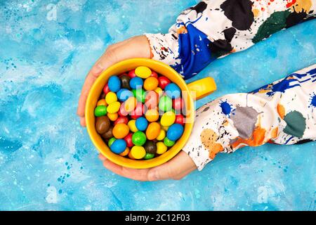 Crop-Ansicht der weiblichen Hände tragen bunte lange Ärmel Sweatshirt mit einem großen Becher voll von bunten Dragee Süßigkeiten auf einem hellen blauen Hintergrund. V Stockfoto