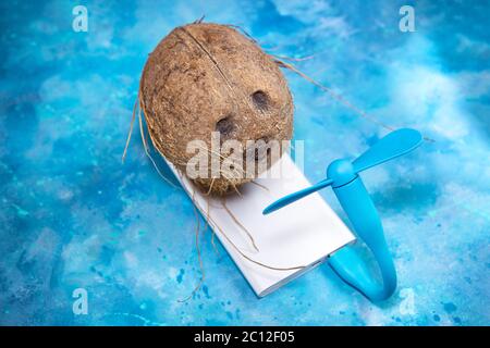 Coconut kühlt vor einem tragbaren Lüfter ab, der von einer mobilen Powerbank auf blauem Hintergrund angetrieben wird. Tropisches Sommer-Hitzewelle-Konzept. Stockfoto