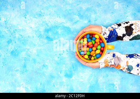 Weibliche Hände in bunten Kleidern halten einen großen Becher mit bunten Schokolade Süßigkeiten vor einem hellen blauen Hintergrund. Draufsicht, Kopierbereich. Stockfoto