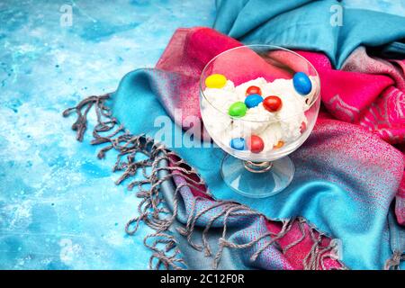 Eis mit bunten Süßigkeiten in einer Glasschale, die auf einem bunten Tuch und hellblauem Hintergrund steht. Helles Dessert-Konzept für den Sommer. Stockfoto