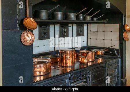 Mehrere Kupfertöpfe unterschiedlicher Größe glänzen im Licht auf dem schwarzen Herd in der Küche des Audley End House nahe Saffron Walden, Essex, England. Ein kleiner Kupferkrug mit Holzgriff steht zwischen den größeren, bedeckten Töpfen. Thomas Howard, 1. Earl of Suffolk, baute das Audley End House zwischen 1605 und 1614 im jakobinischen Stil, einst eines der größten Landhäuser Englands. English Heritage pflegt das Anwesen seit 1984. Stockfoto