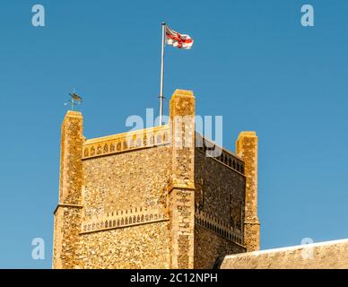 Die Flagge des heiligen Georg – ein rotes Kreuz auf Weiß – weht vom quadratischen Flintstein-Kirchturm der möglicherweise St. Peter und St. Paul in Aldeburgh, Suffolk, England; daneben dreht sich eine Wetterfahne im Wind. Der Turm stammt aus dem 14. Jahrhundert und ist der älteste erhaltene Teil der Kirche, die im frühen 16. Jahrhundert größtenteils im spätgotischen Stil umgebaut wurde. Stockfoto