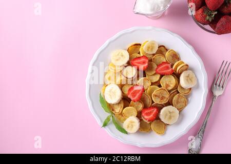 Trendige kleine Pfannkuchen zum Frühstück mit Erdbeere, Banane und saurer Sahne in weißem Teller auf rosa Hintergrund. Draufsicht. Platz für Text oder Design kopieren Stockfoto