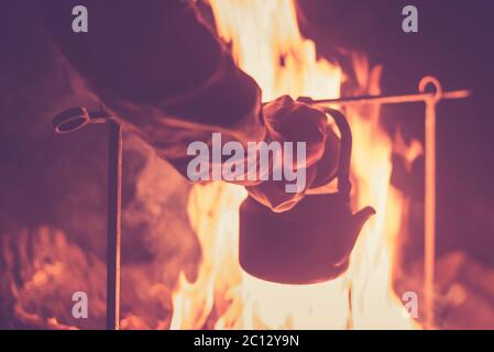 Hand eines Mannes, der den Wasserkocher nahm, der nachts an einem hellen Feuer im Freien erhitzt wurde, getöntes Foto Stockfoto