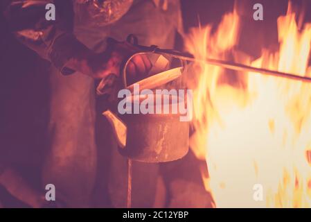 Hand eines Mannes, der den Wasserkocher nahm, der nachts an einem hellen Feuer im Freien erhitzt wurde, getöntes Foto Stockfoto