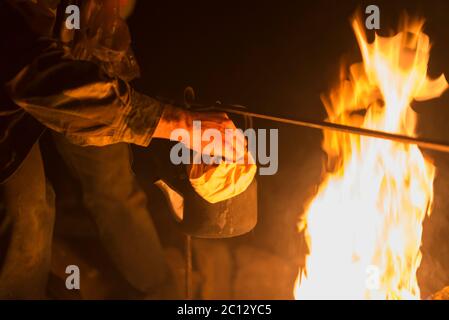 Hand eines Mannes, der den Wasserkocher nahm, der nachts draußen an einem hellen Feuer erhitzt wurde Stockfoto