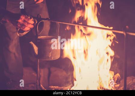 Hand eines Mannes, der den Wasserkocher nahm, der nachts an einem hellen Feuer im Freien erhitzt wurde, getöntes Foto Stockfoto