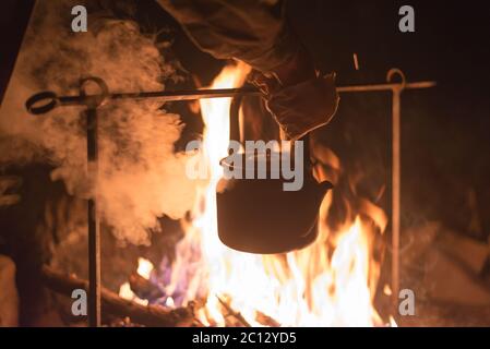 Hand eines Mannes, der den Wasserkocher nahm, der nachts draußen an einem hellen Feuer erhitzt wurde Stockfoto