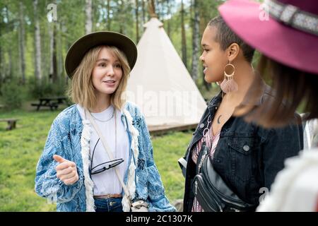 Ziemlich blonde Mädchen im Hut im Chat mit Freunden auf dem Festival-Campingplatz, sie planen Freizeit in der Landschaft Stockfoto