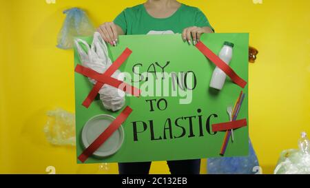 Nicht erkennbare Frau Freiwillige in T-Shirt mit Recycling-Logo hält protestierende Nachricht Poster Say No to Plastic. Hintergrund mit Zellophanbeutel, Flaschen. Umweltmüll Plastikverschmutzung Stockfoto