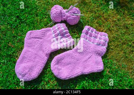 Ein Paar rosa Wollsocken und ein Garnball mit einem Satz Nadeln auf einem grünen Gras Hintergrund. Handgefertigte Kunstsocken mit weißen Streifen, weicher Stoff Stockfoto