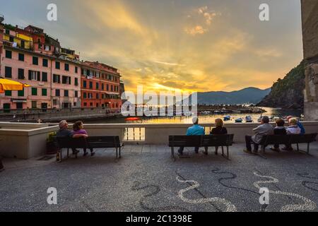 Touristen genießen einen bunten Sonnenuntergang über dem ligurischen Meer, während sie auf einer Terrasse in der Nähe des kleinen Hafens im Cinque Terre Dorf Vernazza, Italien sitzen Stockfoto