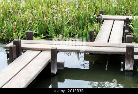 Eine japanische Zickzack-Brücke über einen kleinen Teich durch üppige Gräser und Blumen Stockfoto