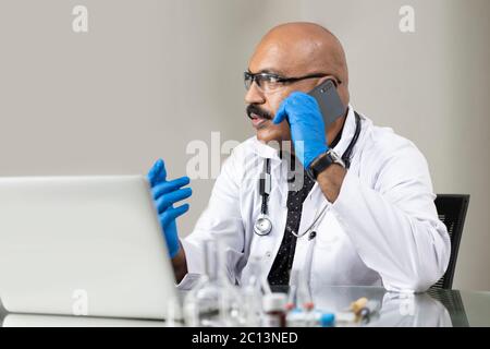 Indische männliche Arzt unterstützt Patienten online mit Telefon und Computer Während dieser Pandemie Stockfoto