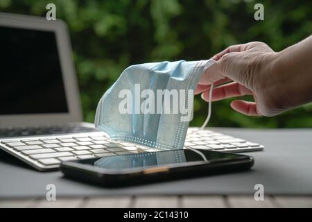 Frau hält medizinische Gesichtsmaske mit Computer Laptop Tastatur und Handy in Sicht. Arbeiten zu Hause während covid-19 Konzept. Stockfoto